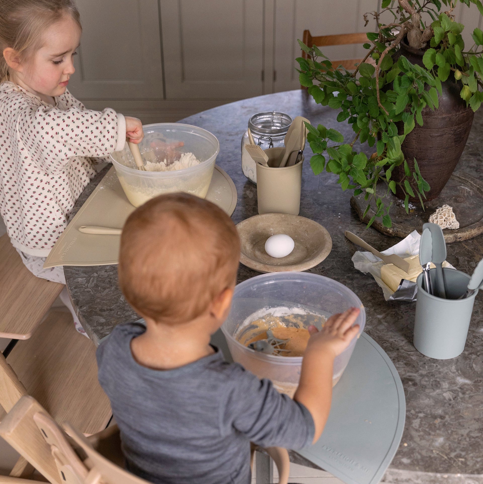 To børn sidder og laver mad på højstole med hver deres skål. De bruger silikone køkkenredskaber fra Everleigh and Me til at bage muffins. Køkkenredskaber passer perfekt til de små barnehænder og inviterer børnene til aktivt at deltage i bagningen.