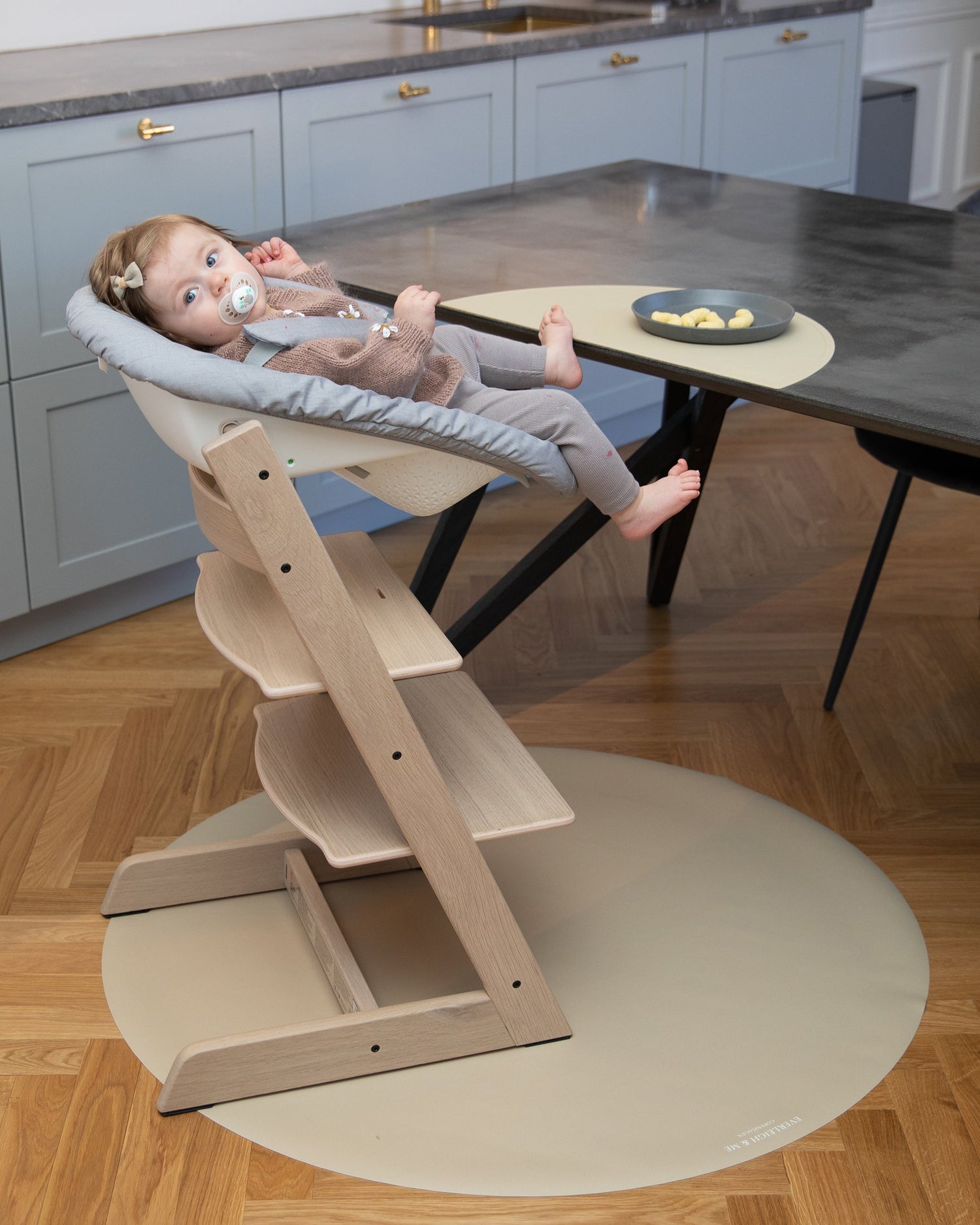 En baby sidder på en stokke træ højstol og spiser efter baby-led weaning-metoden, mens et stoleunderlag i farven Clay fra danske børnemærke Everleigh &amp; Me beskytter gulvet mod spild. Underlaget smukt matcher gulvets farvetone. underlag gulv, baby stole, underlag kontorstol, plastmåtte