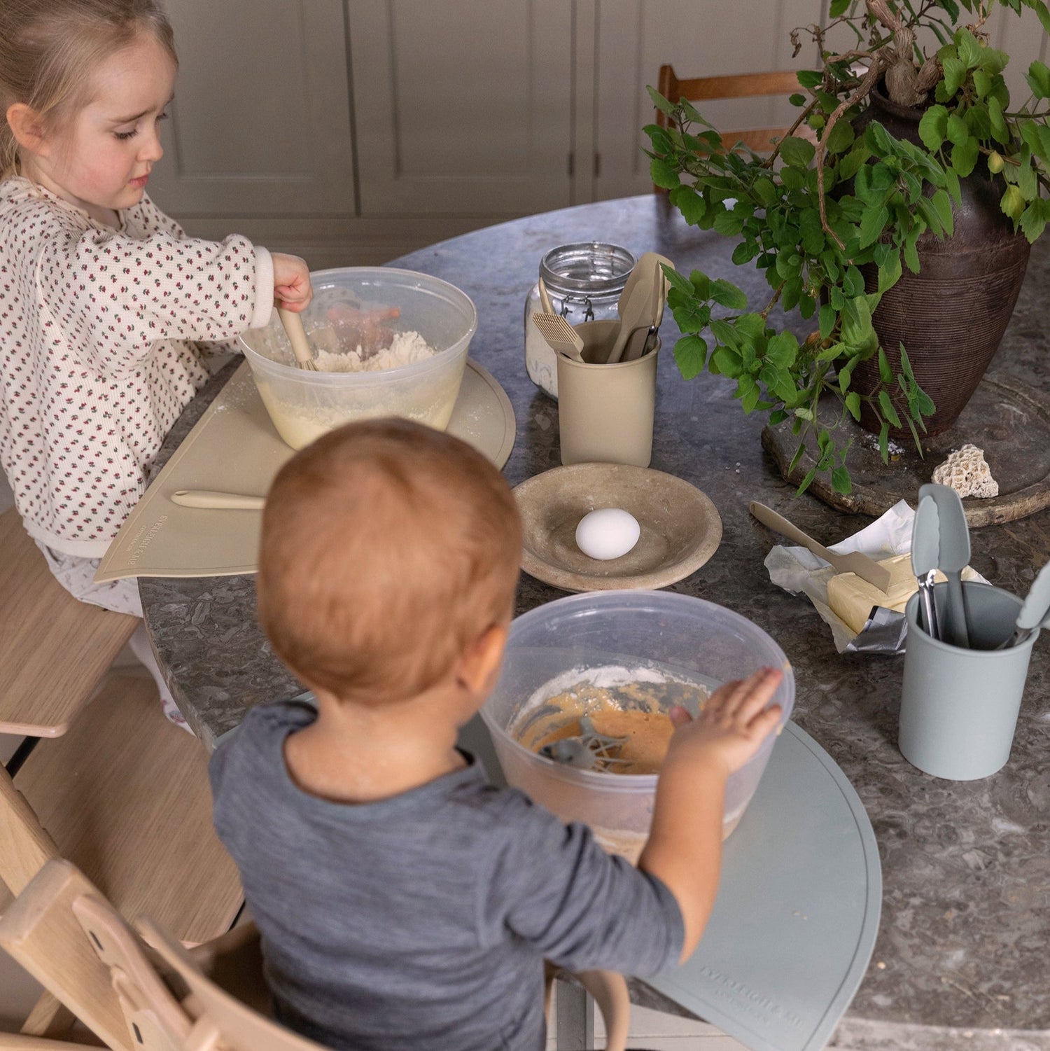 To børn sidder og laver mad på højstole med hver deres skål. De bruger silikone køkkenredskaber fra Everleigh and Me til at bage muffins. Køkkenredskaber passer perfekt til de små barnehænder og inviterer børnene til aktivt at deltage i bagningen.