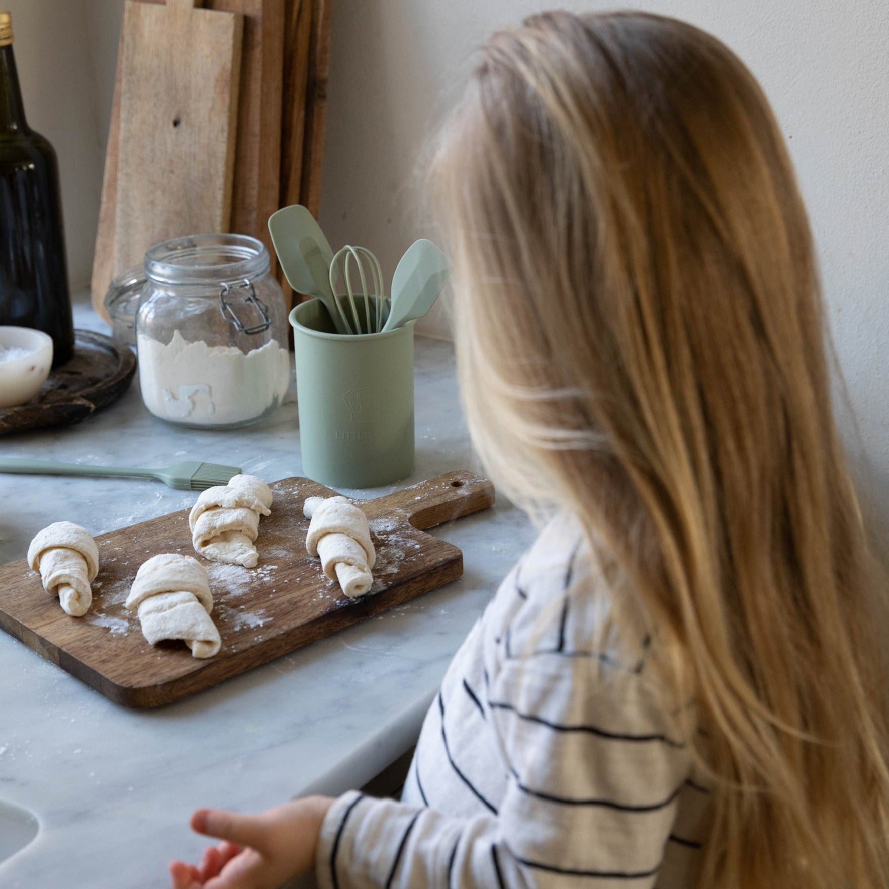 Barn i gang med madlavning ved brug af Olive køkkensæt fra Everleigh &amp; Me. De børnevenlige redskaber i en varm olivengrøn farve gør madlavningen sjov og lærerig i et stilfuldt køkken.